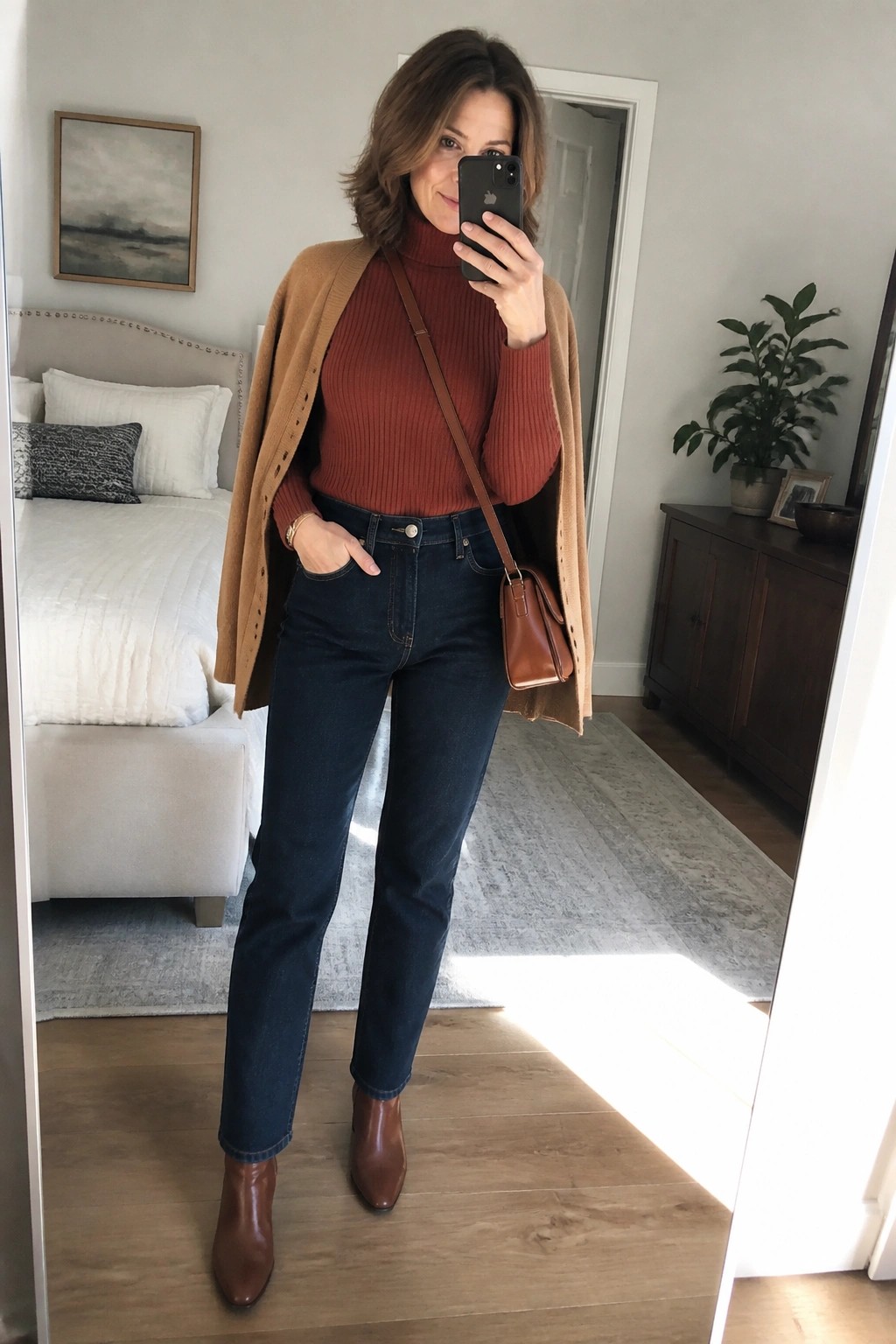A woman wearing a rust turtleneck sweater under an open camel cardigan, paired with dark straight leg jeans, brown ankle boots, and a matching brown crossbody bag, shown in a full length mirror selfie.