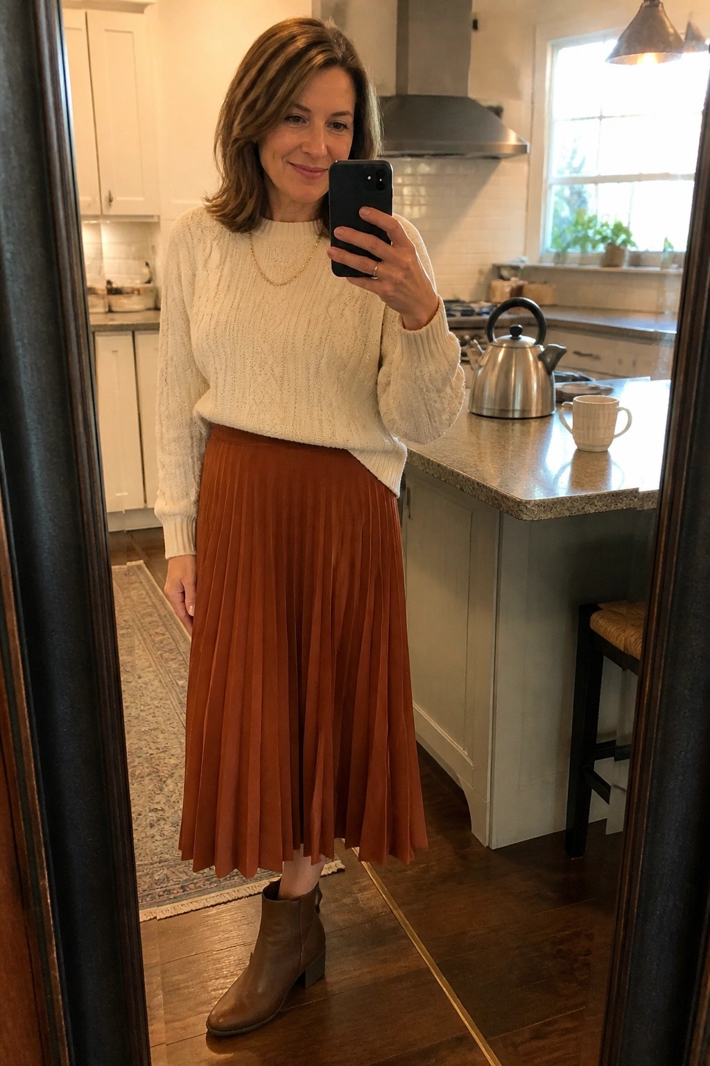 Woman in cream sweater and rust pleated skirt taking mirror selfie in kitchen