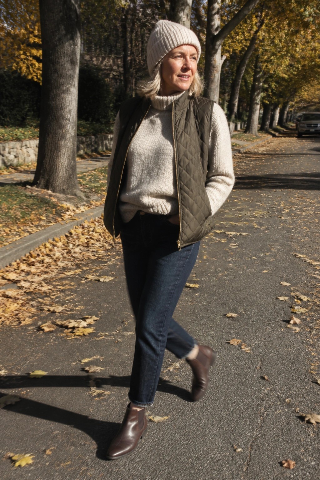 Woman in cream sweater, green quilted vest, and beanie walks on autumn street