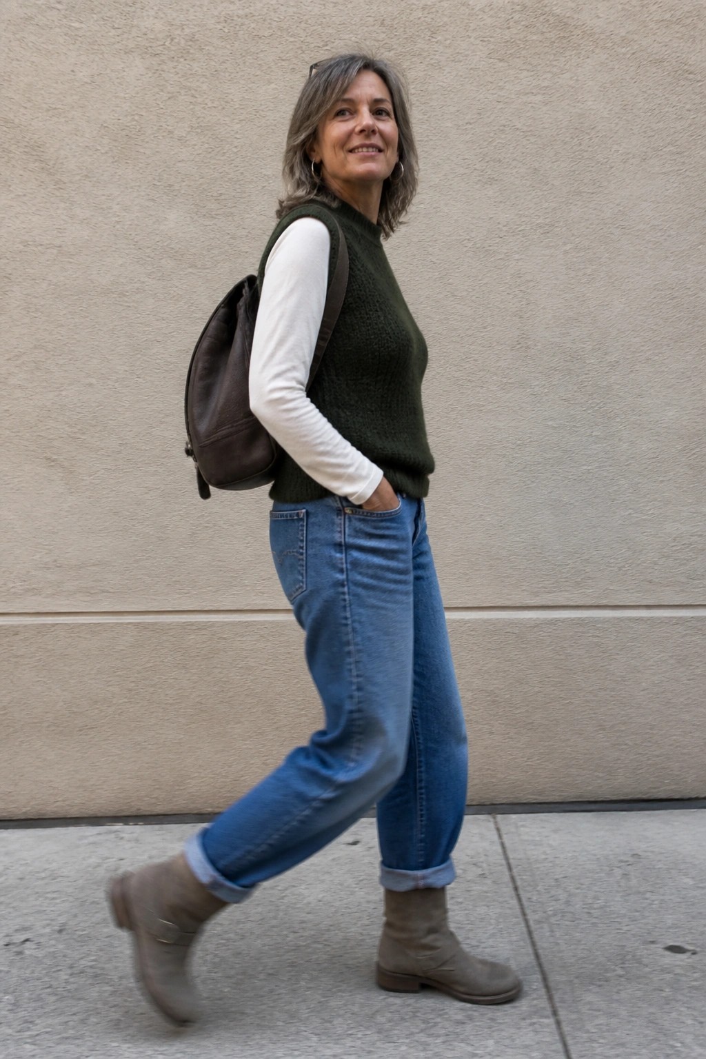 Woman over 50 wearing a green knit vest over a white long-sleeve shirt with blue jeans and taupe suede ankle boots carrying a brown leather backpack.