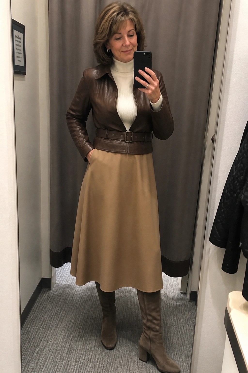 Woman in brown leather jacket and tan skirt takes mirror selfie with phone