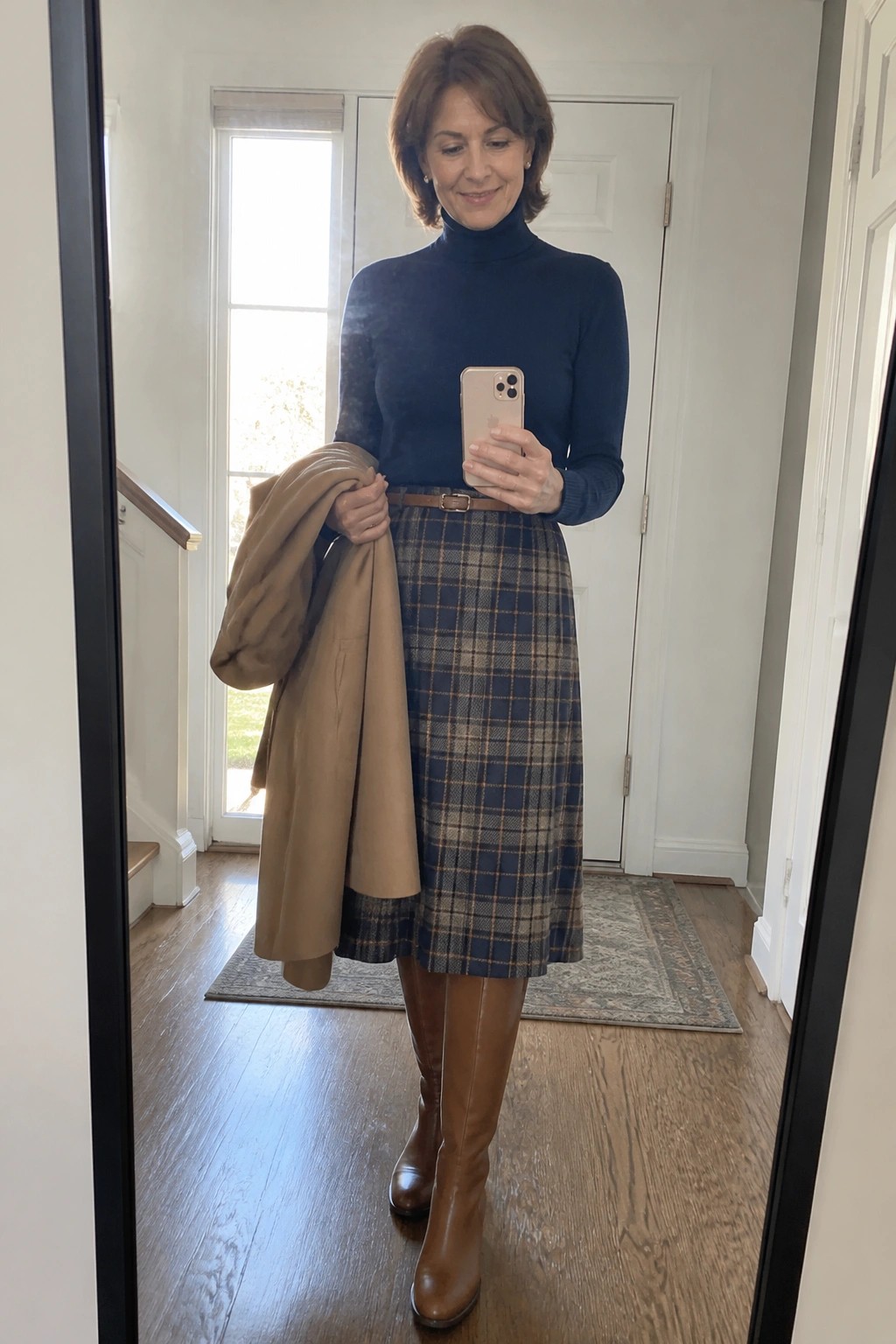 Woman over 50 wearing navy turtleneck sweater tucked into blue brown plaid skirt with brown belt and tall brown leather boots while holding tan coat in hallway mirror selfie