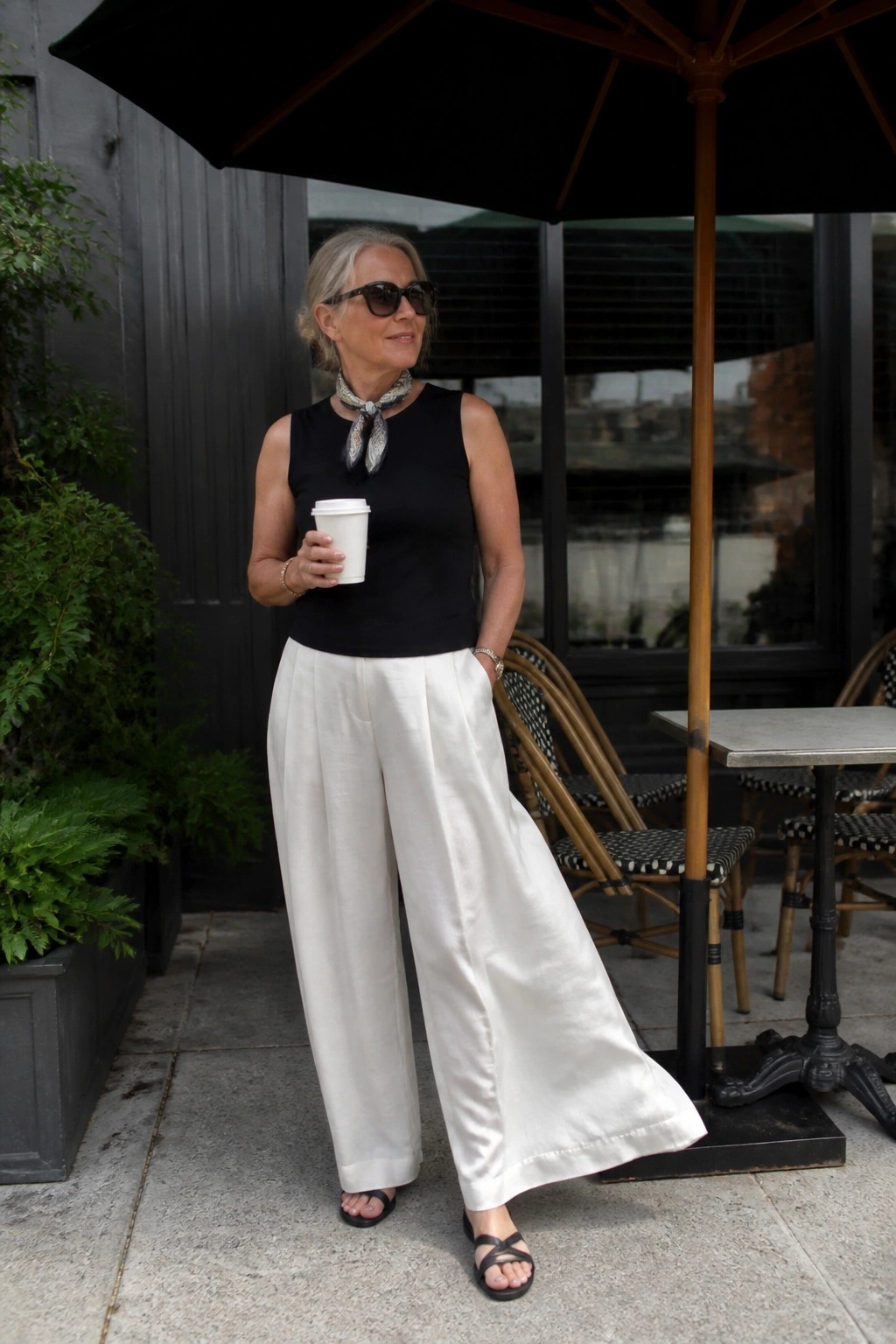 Woman in black top and white wide-leg pants holding coffee at outdoor cafe