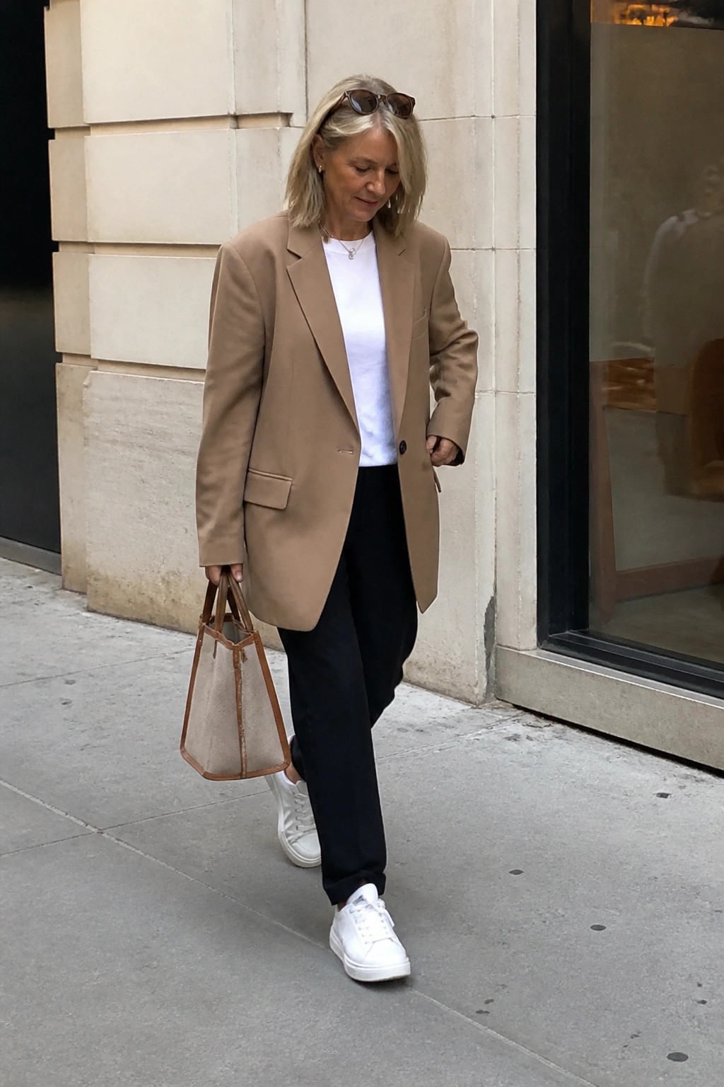 Woman in tan blazer, white tee, black pants, and sneakers carrying mesh tote on sidewalk