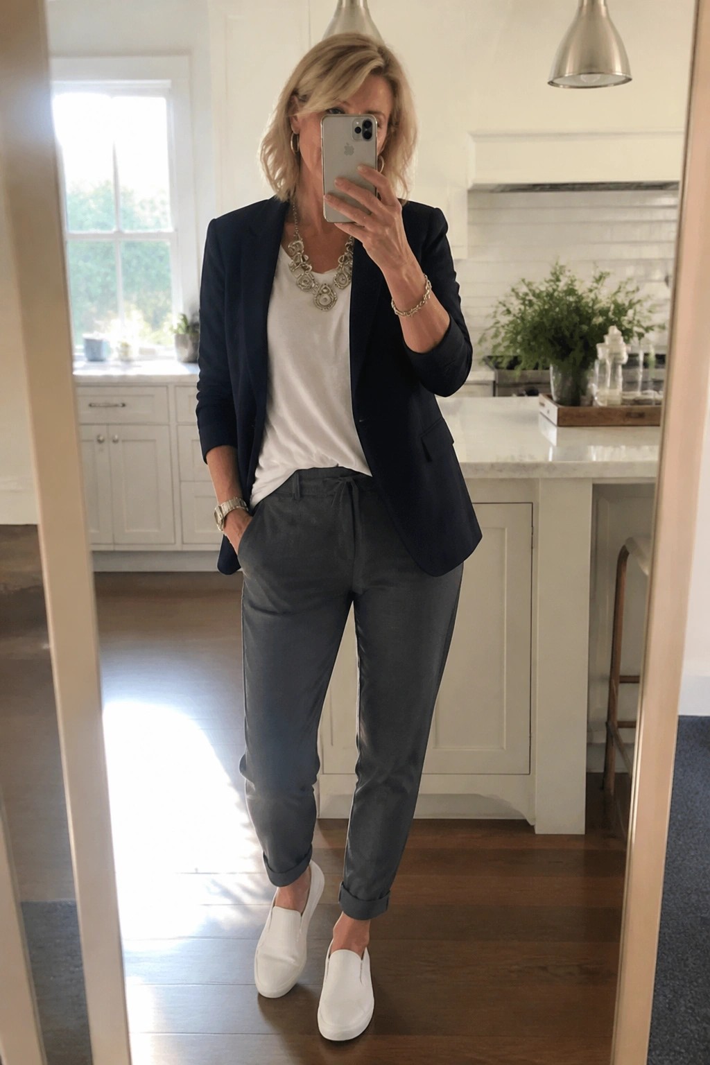 Blonde woman in navy blazer, white top, and gray pants taking kitchen mirror selfie