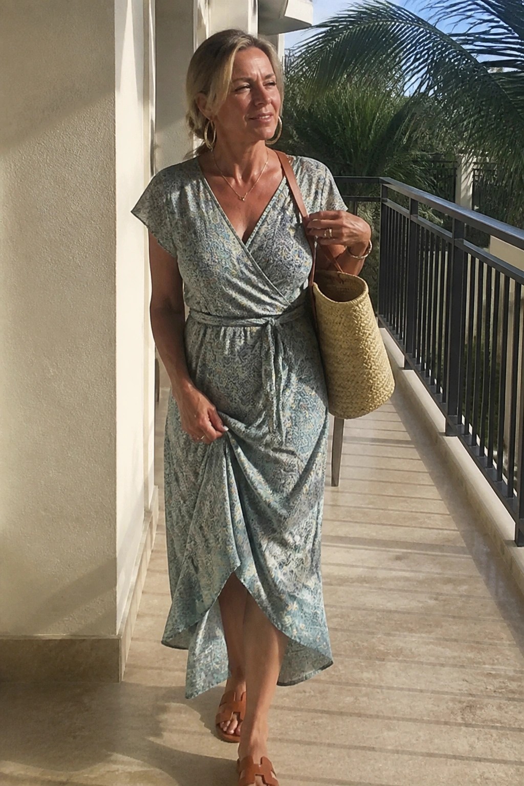 Blonde woman in light blue floral wrap dress with woven straw bag on sunny balcony