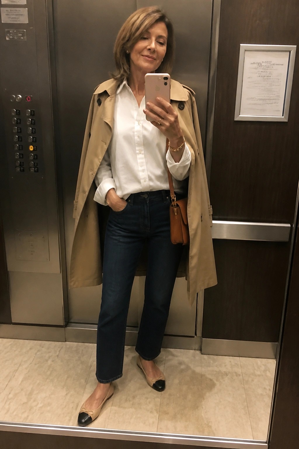 Woman wearing an open beige trench coat over a white button down shirt tucked into straight leg blue jeans with beige and black flats and a brown leather crossbody bag.