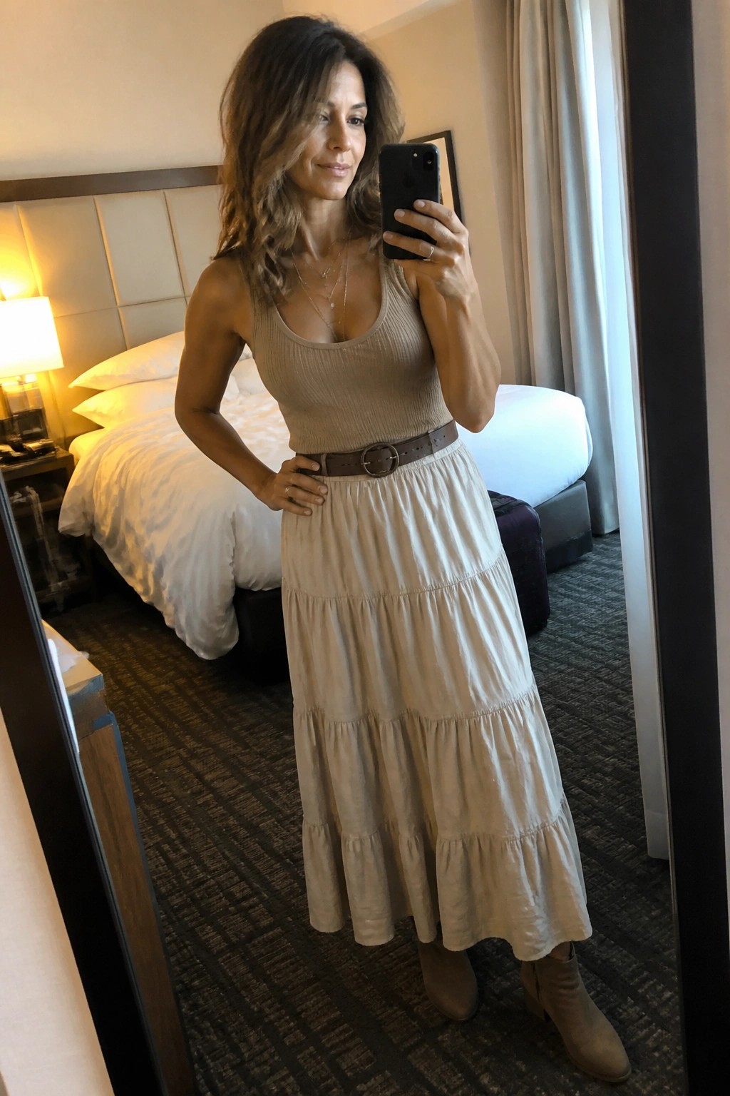 Woman in beige tank top and tiered skirt taking mirror selfie in hotel room