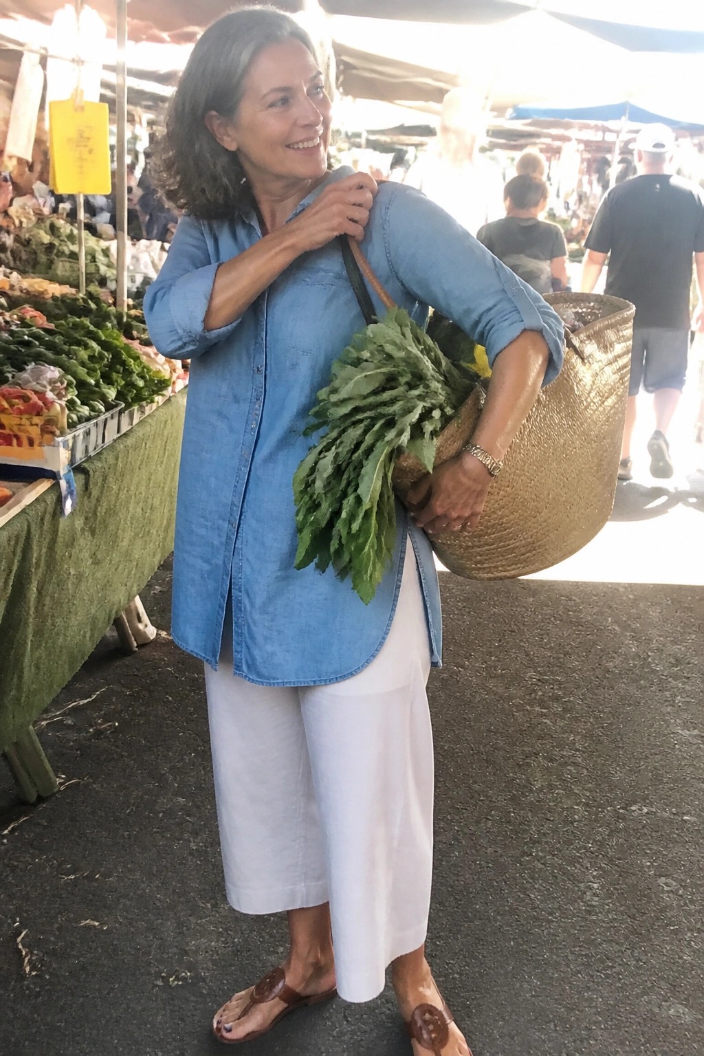 Smiling gray-haired woman carries woven basket of greens at outdoor market