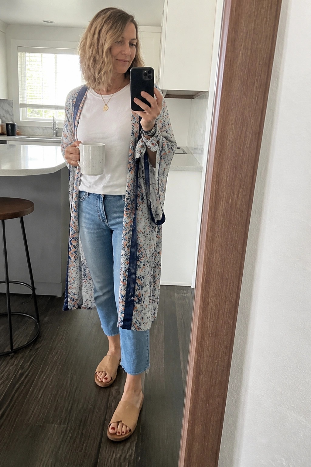 Woman over 50 in a long floral duster jacket layered over a white crewneck tee and cropped blue jeans with tan slide sandals plus layered necklaces and bracelets