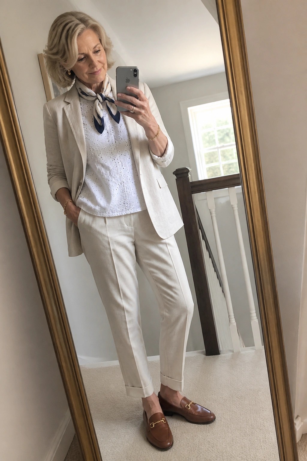 Woman in white blazer, eyelet blouse, and pants taking mirror selfie with iPhone.