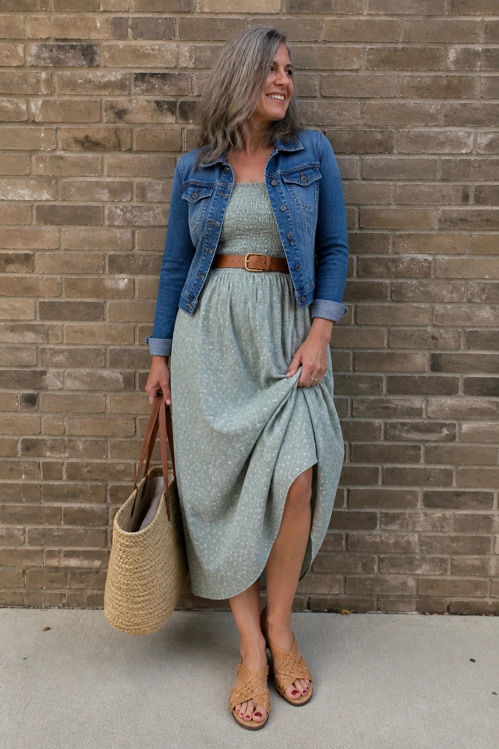 Woman over 50 wearing a sage green smocked midi dress with white floral print under a blue denim jacket belted at the waist with brown leather, paired with woven leather sandals and a large straw tote bag.