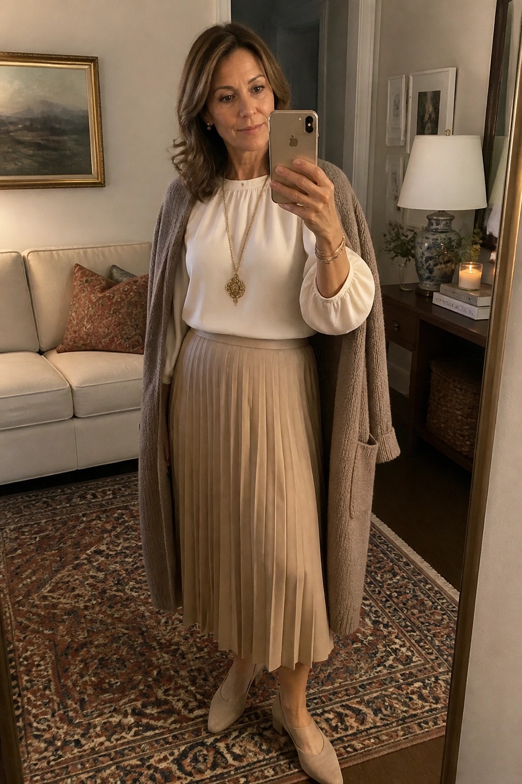 Woman over 50 in mirror selfie wearing white blouse with gathered sleeves, long beige pleated skirt, taupe draped cardigan, gold pendant necklace, and matching beige shoes.