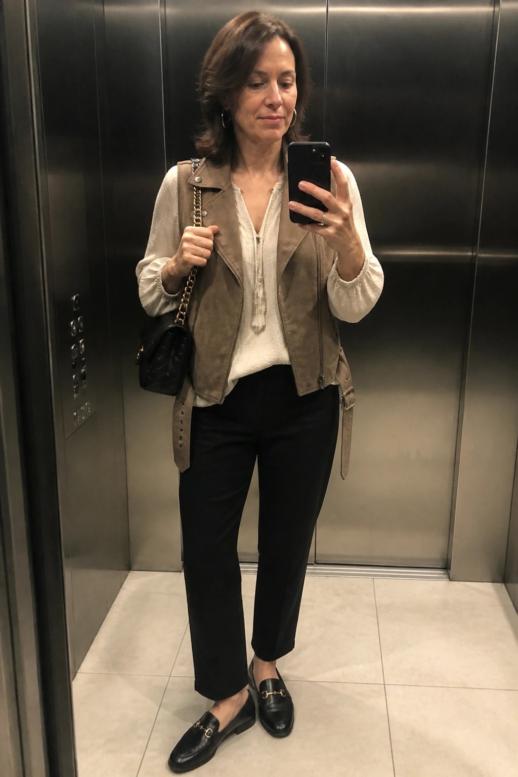 Woman in elevator taking selfie, wearing beige suede vest, white blouse, black pants and loafers.