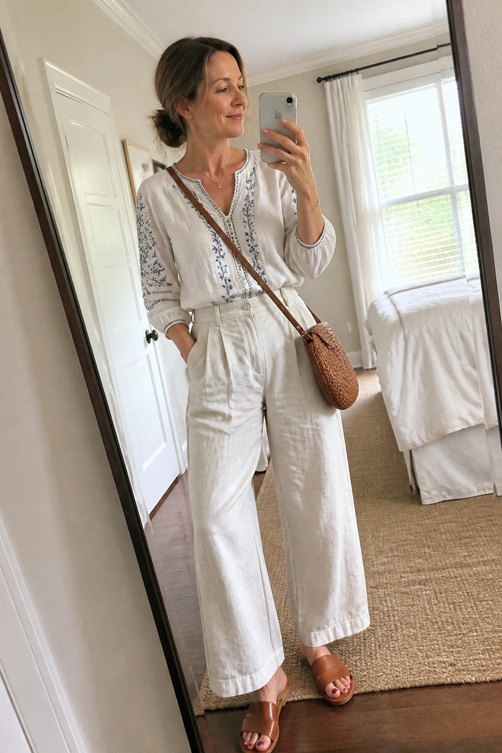 Woman wearing white linen wide leg pants with matching embroidered blouse and brown slide sandals holding a phone for a mirror selfie indoors