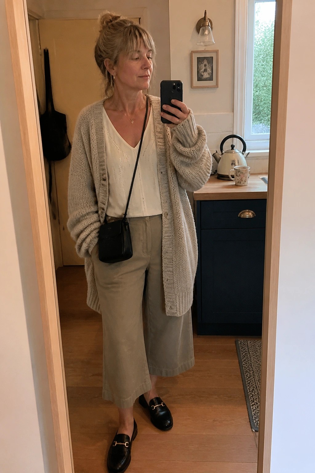 Woman in beige knit cardigan and wide pants taking mirror selfie in kitchen
