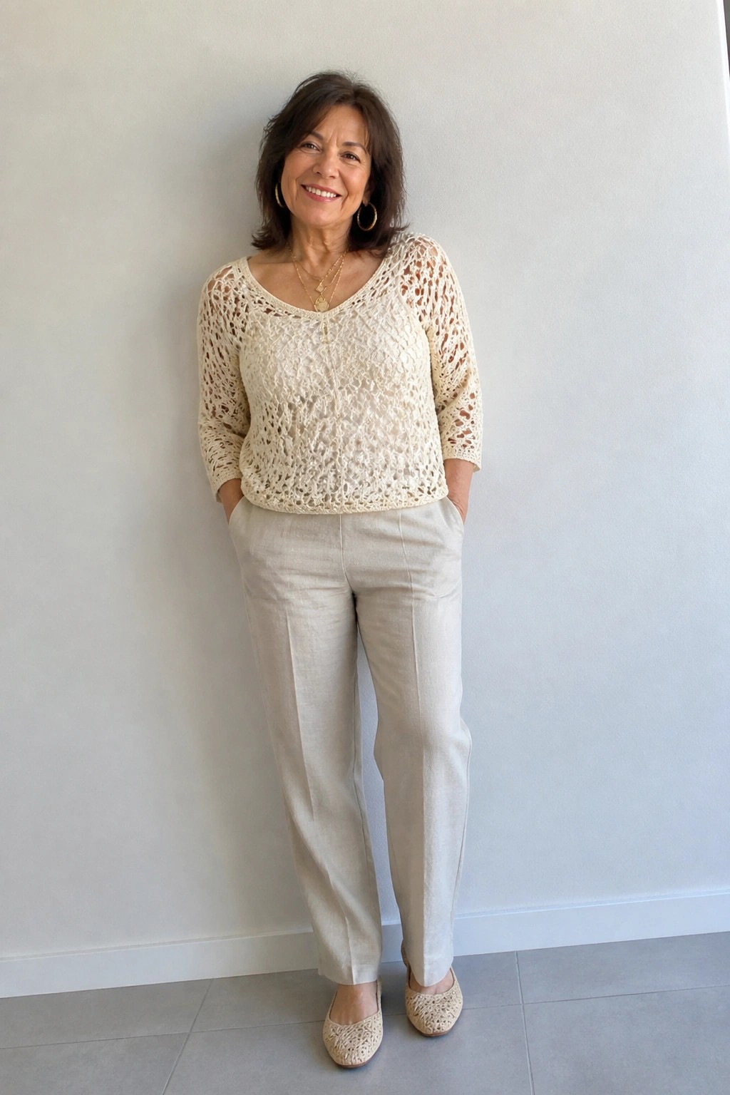 Smiling woman in cream crocheted top and beige pants with hands in pockets.