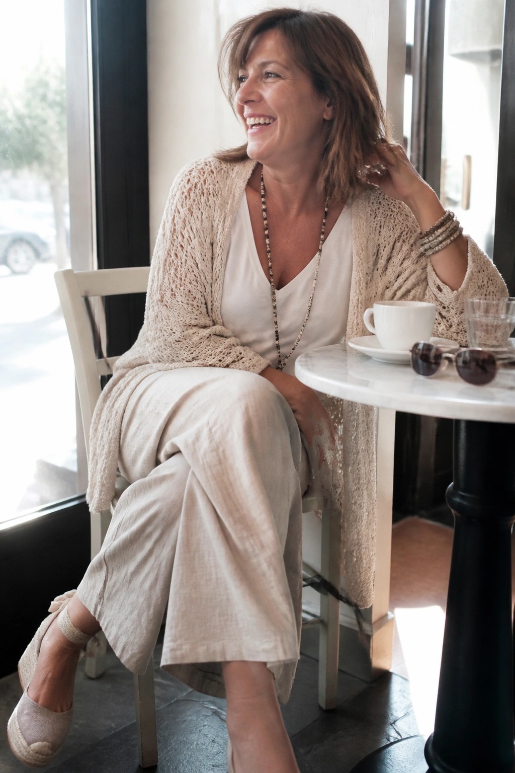 Smiling woman in beige knit cardigan and linen pants at marble cafe table with coffee.