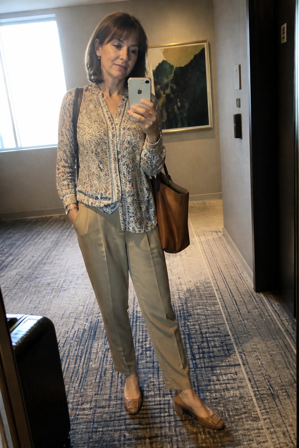 Woman in floral blouse and beige pants taking mirror selfie in hotel hallway