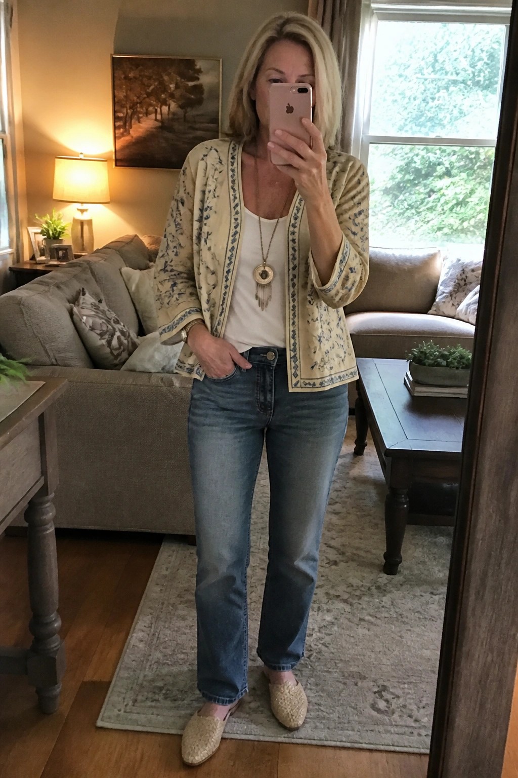 Blonde woman in cream patterned jacket and jeans taking mirror selfie in living room