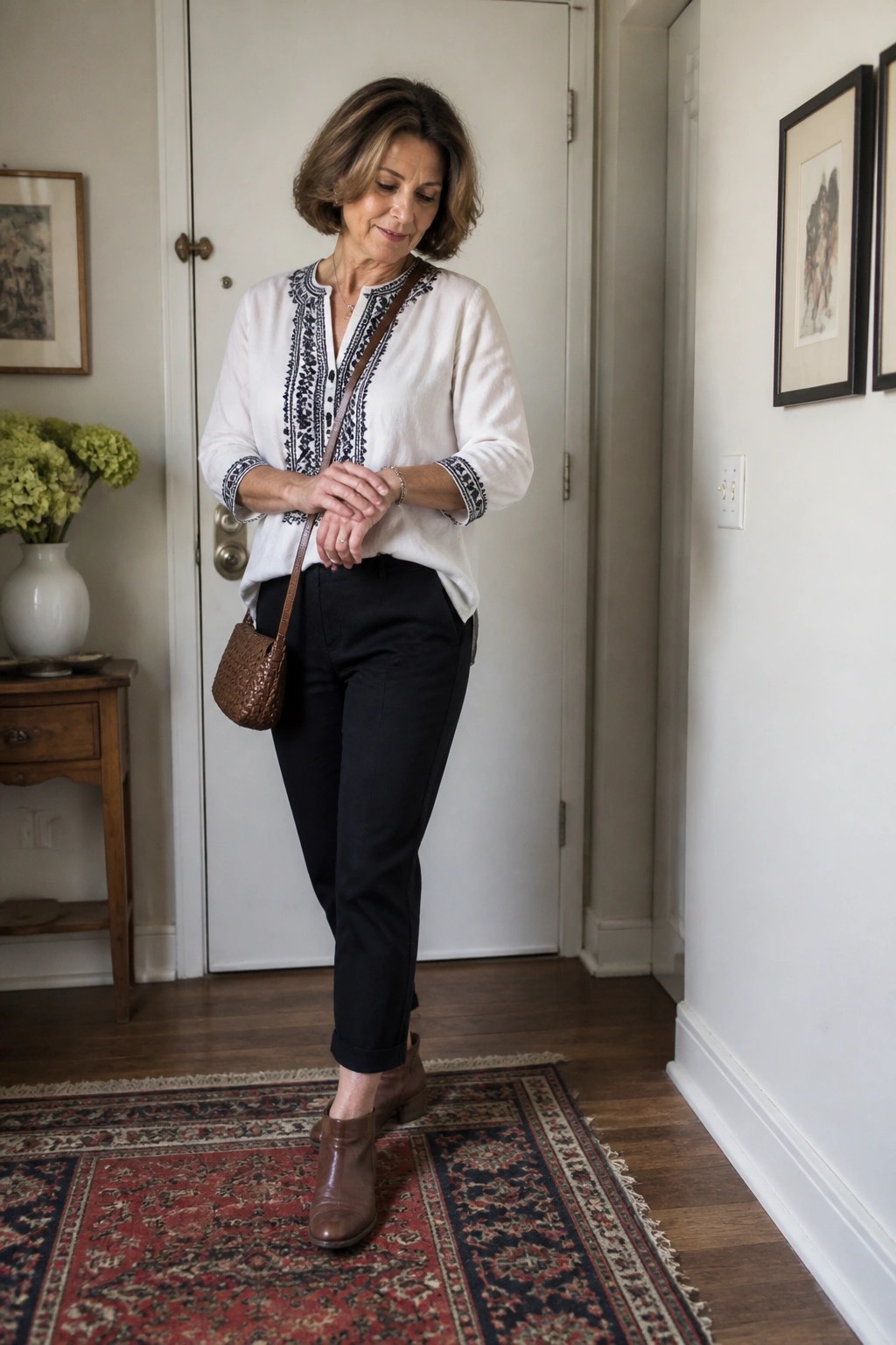 Woman over 50 in a white embroidered blouse tucked into black pants with brown ankle boots and a woven brown crossbody bag standing on a patterned rug.