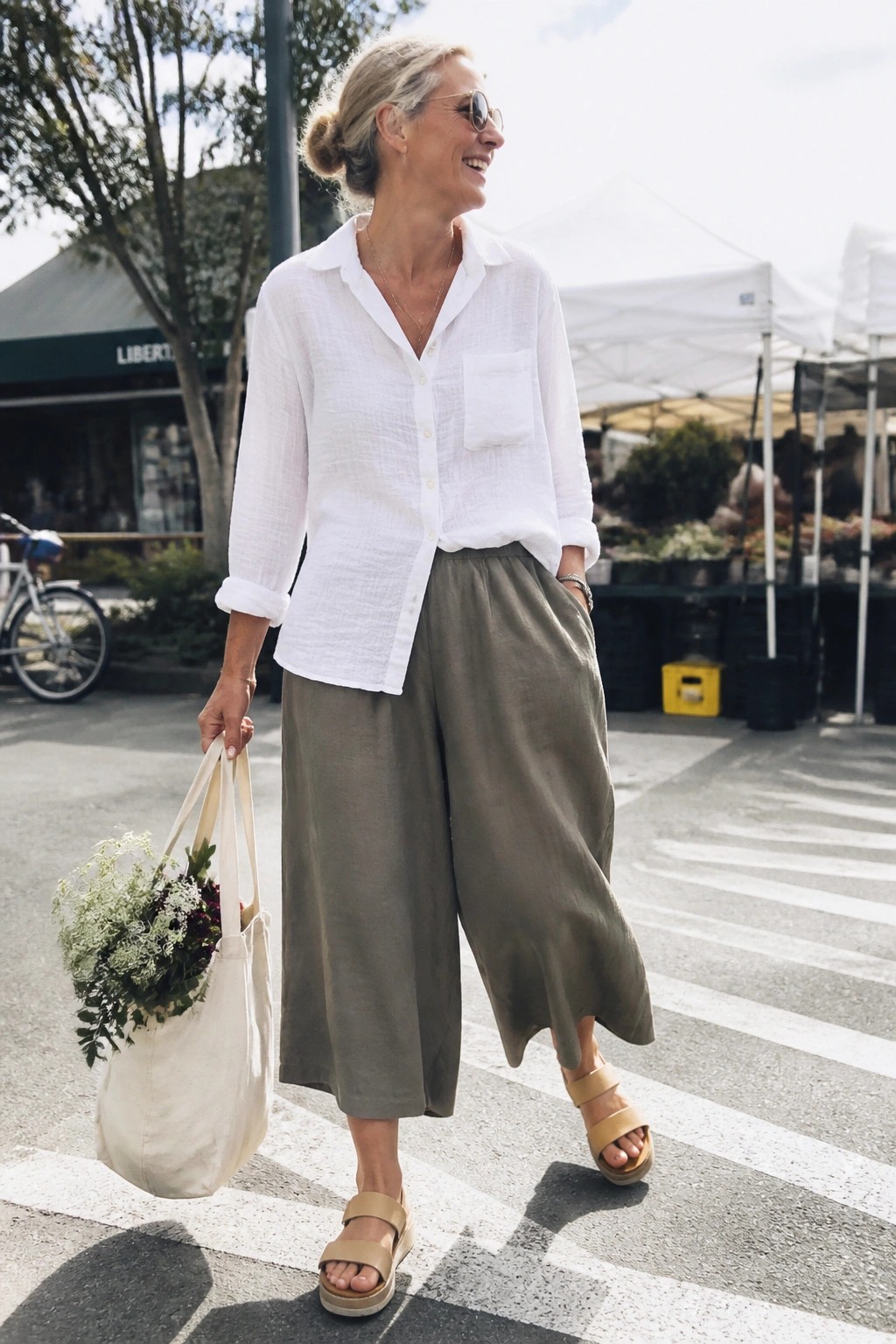 Woman over 50 wearing a white linen button-up shirt with rolled sleeves tucked into olive green wide-leg cropped linen pants, paired with beige platform sandals and carrying a cream tote bag filled with flowers, layered gold necklaces visible at the neckline.
