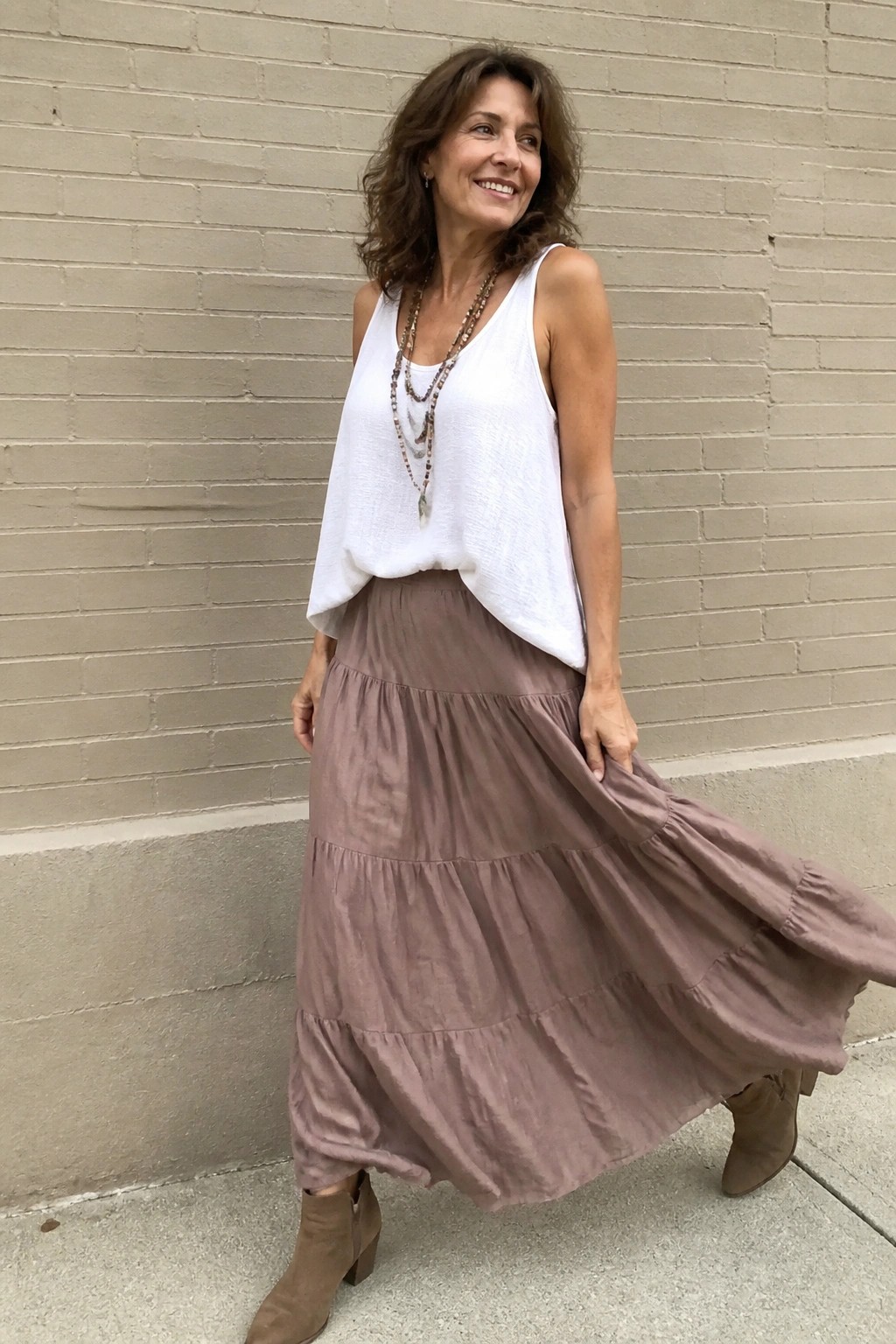 Woman over 50 wearing a loose white tank top tucked into a long taupe tiered maxi skirt with brown suede ankle boots and a layered beaded necklace standing against a brick wall.