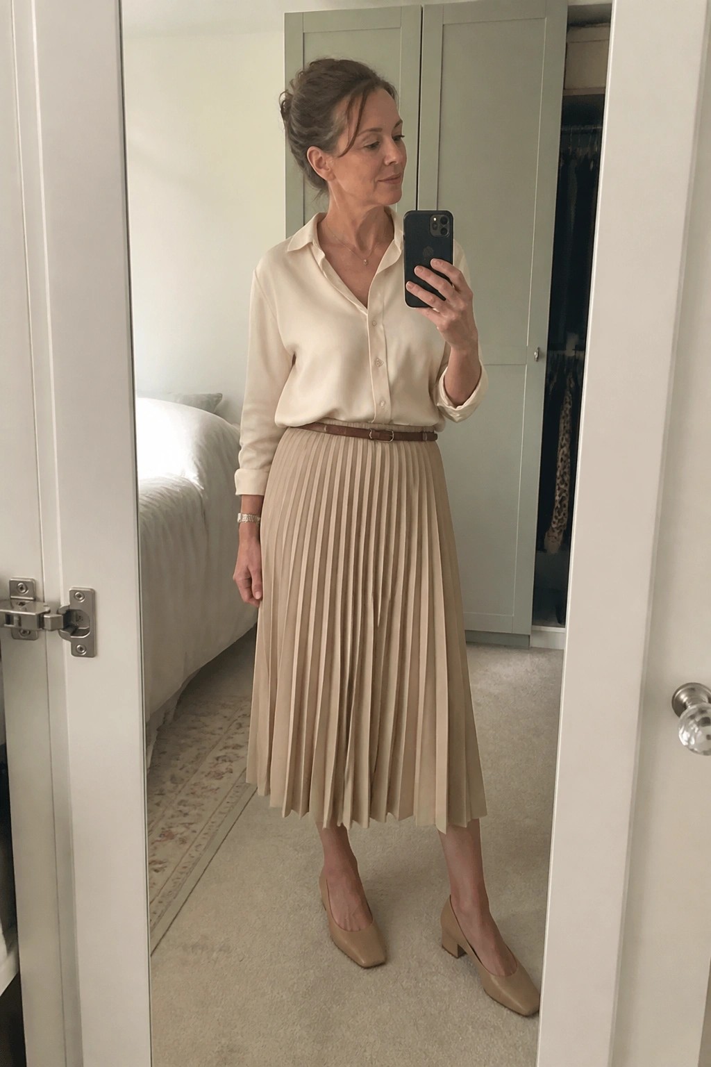 Woman in cream blouse and beige pleated skirt taking mirror selfie with phone