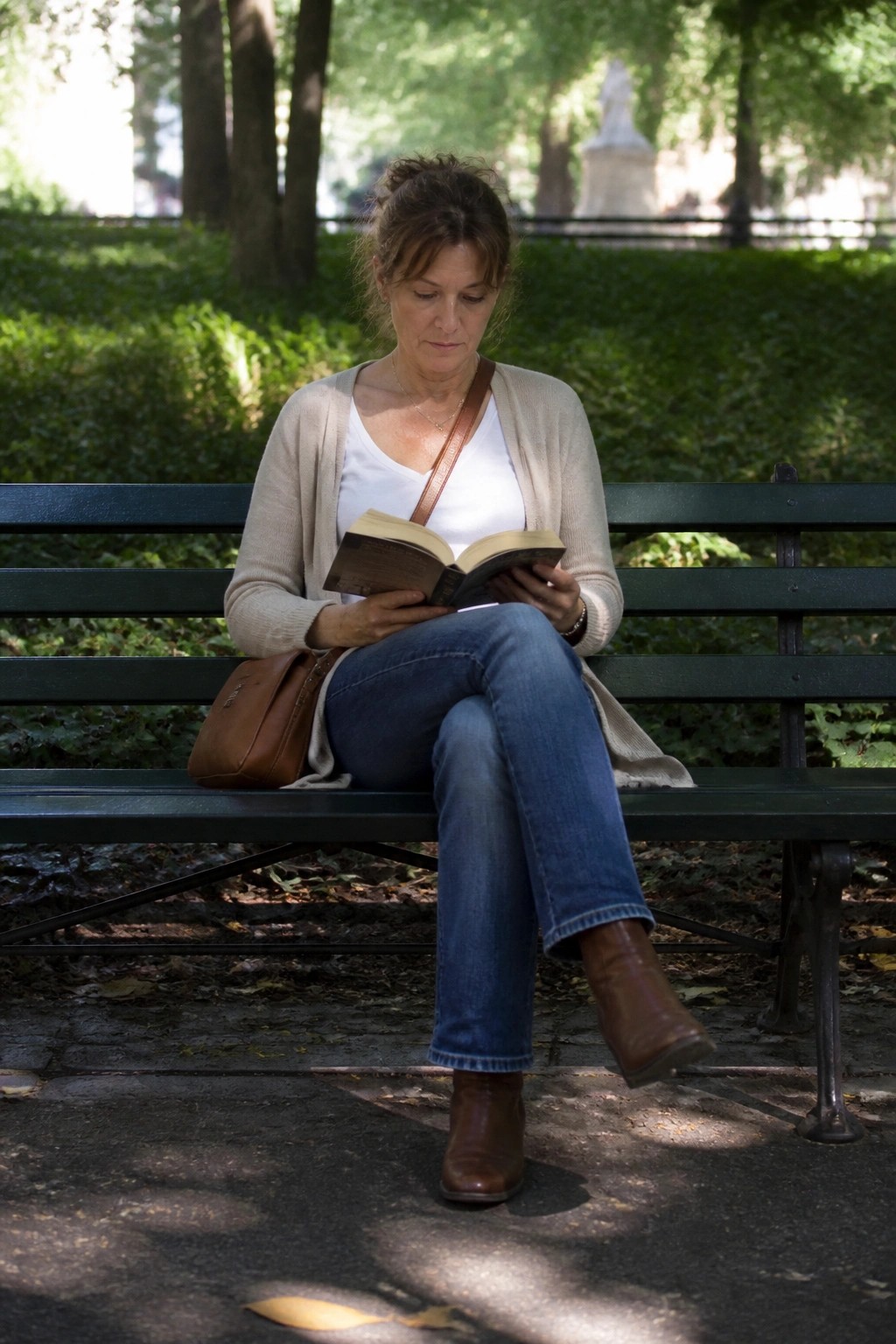Woman in beige cardigan reading book on green park bench with brown bag.