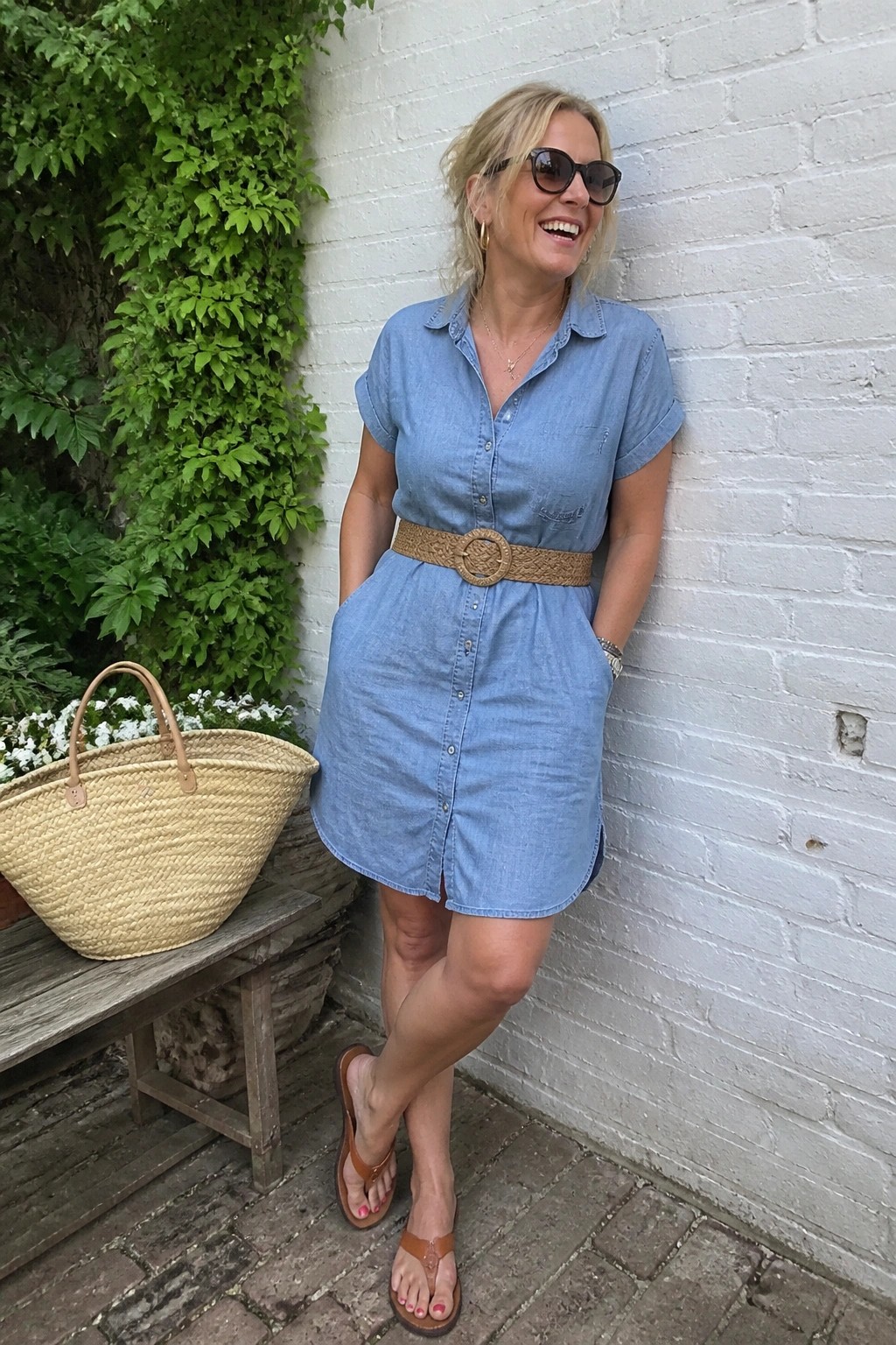 Woman in light blue denim shirt dress belted with woven brown belt standing near brick wall with straw tote and brown flip flops
