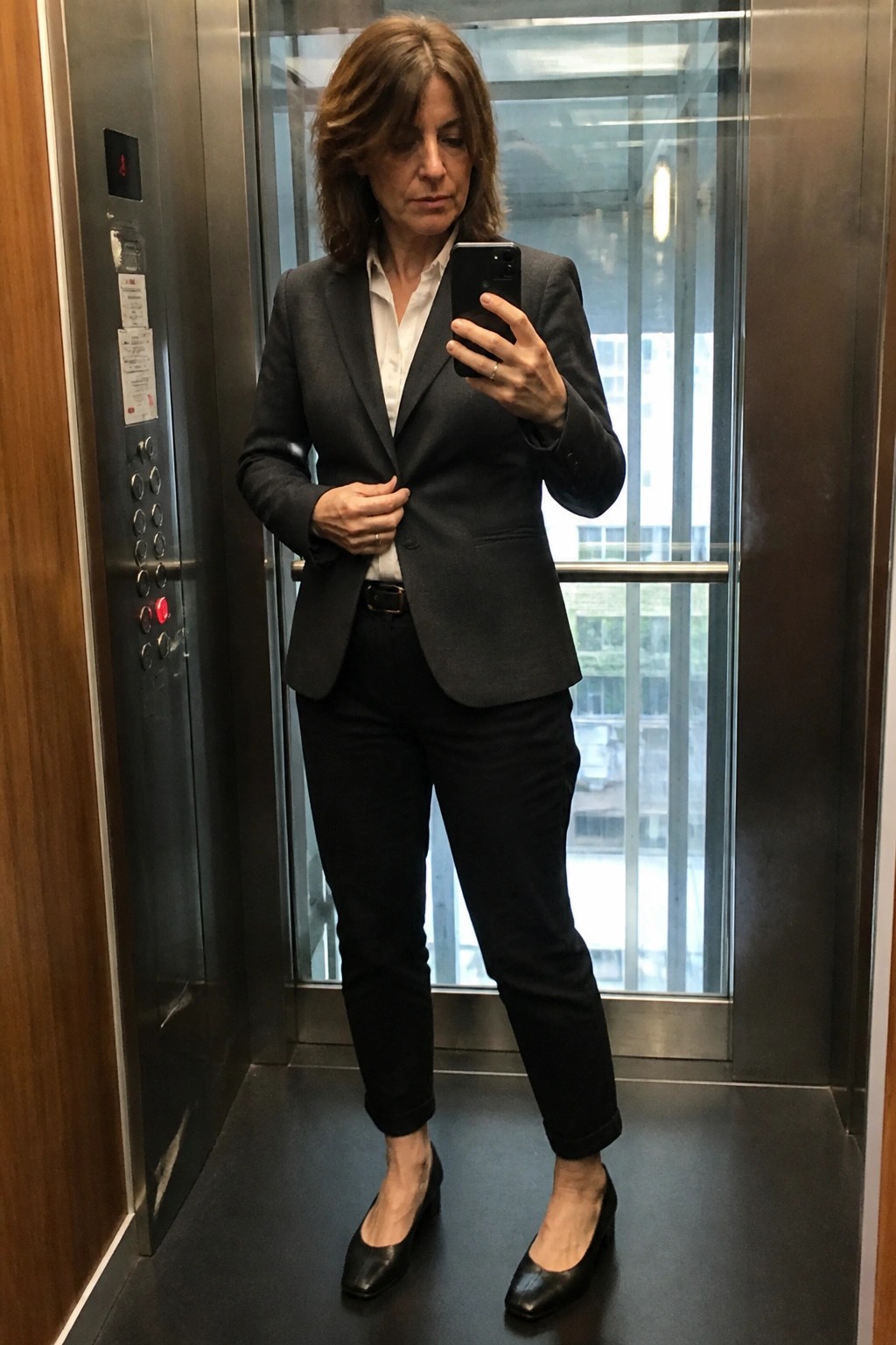 Woman in gray blazer taking selfie with phone in elevator.