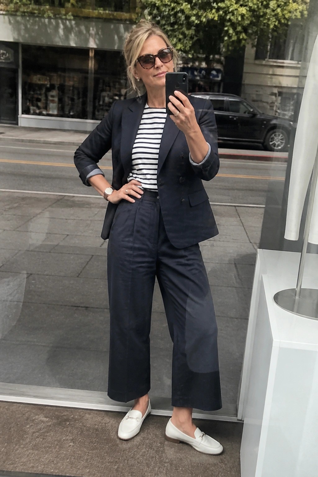 Woman in navy blazer and striped top takes selfie on city sidewalk