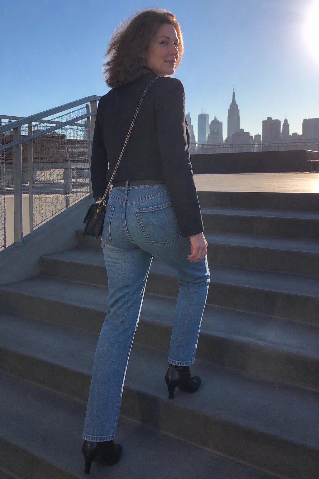 Woman in black blazer and blue jeans on stairs overlooking city skyline