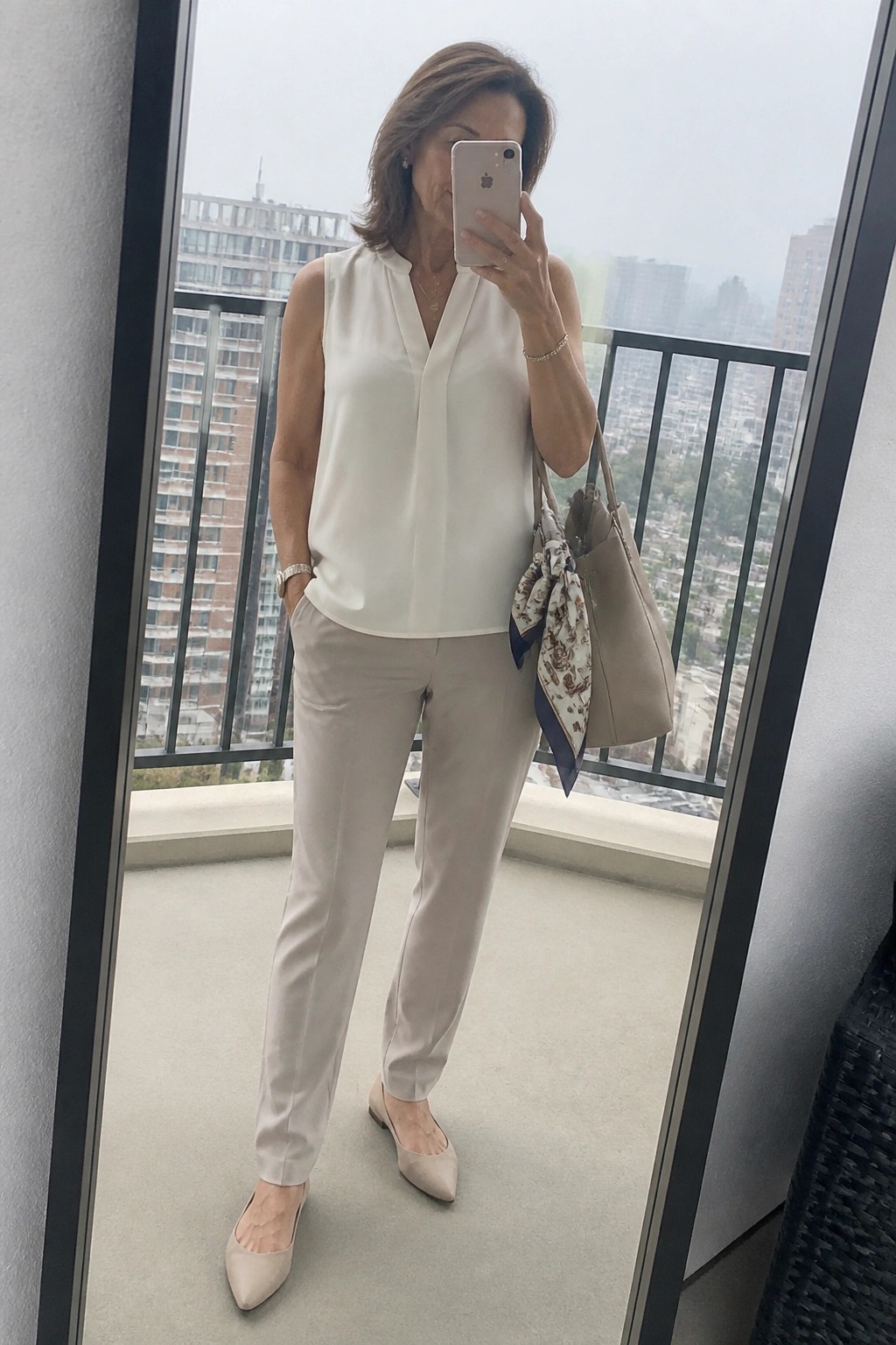 Woman in white sleeveless blouse and beige pants taking balcony selfie with handbag