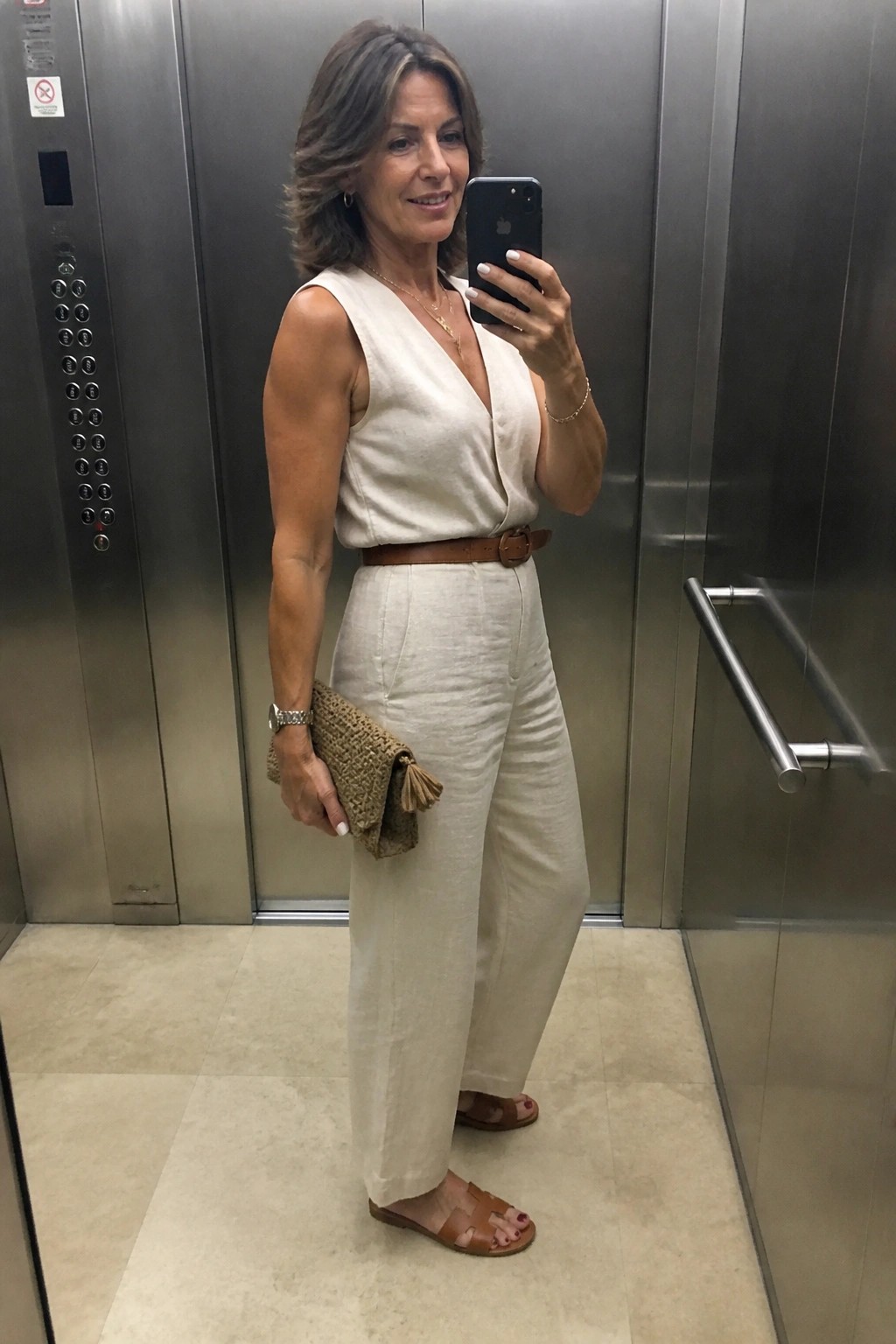 Woman in beige linen jumpsuit taking selfie in elevator with woven clutch