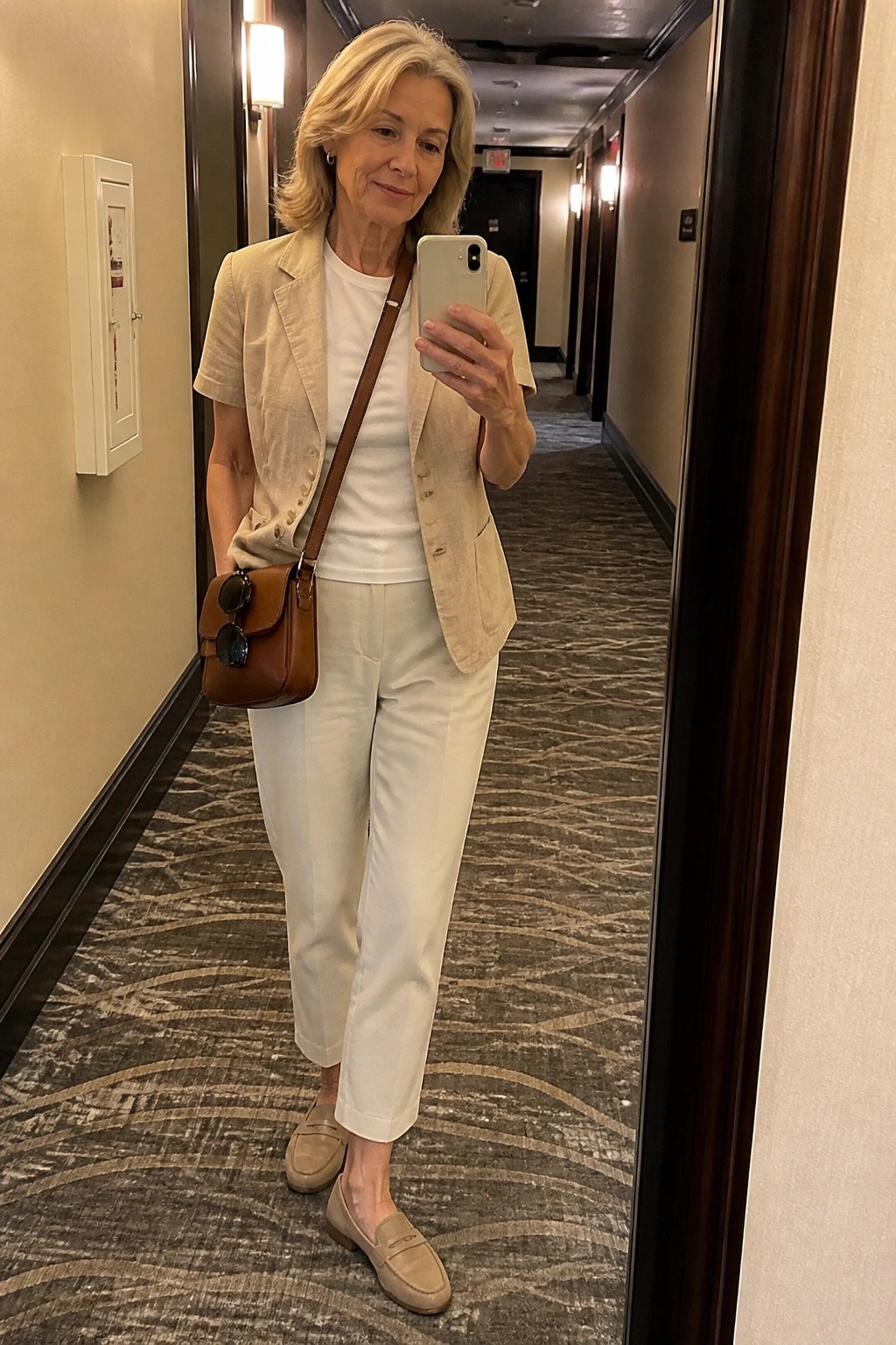 Woman in beige blazer and white pants taking mirror selfie in hallway