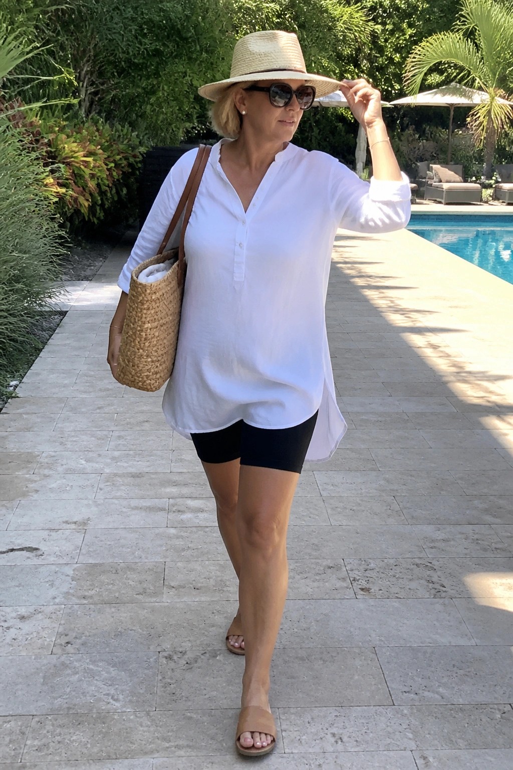Woman in white tunic and black shorts adjusting straw hat by pool with tote bag