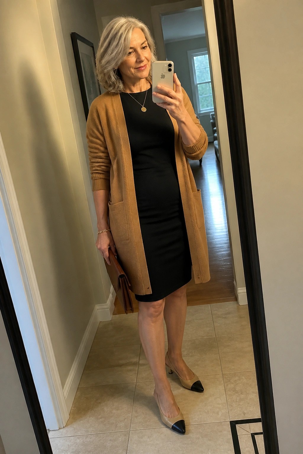 Woman with gray hair in black dress and tan cardigan taking mirror selfie