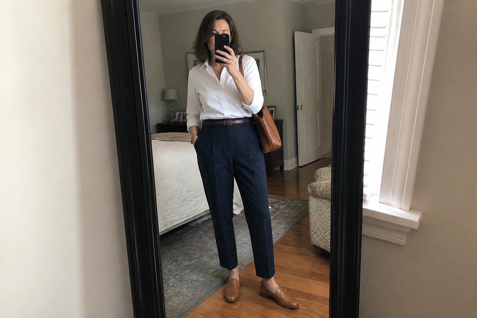 Woman taking a mirror selfie in a white button down shirt tucked into navy pleated trousers with a brown belt brown loafers and a matching crossbody bag.