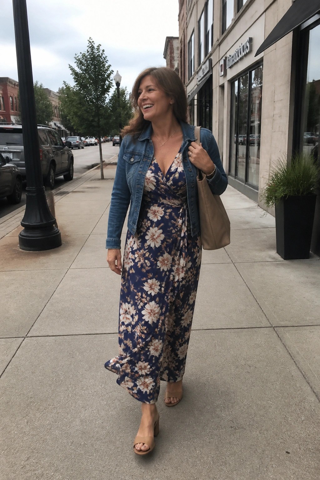 Woman in blue floral dress and denim jacket walking on sidewalk with beige handbag