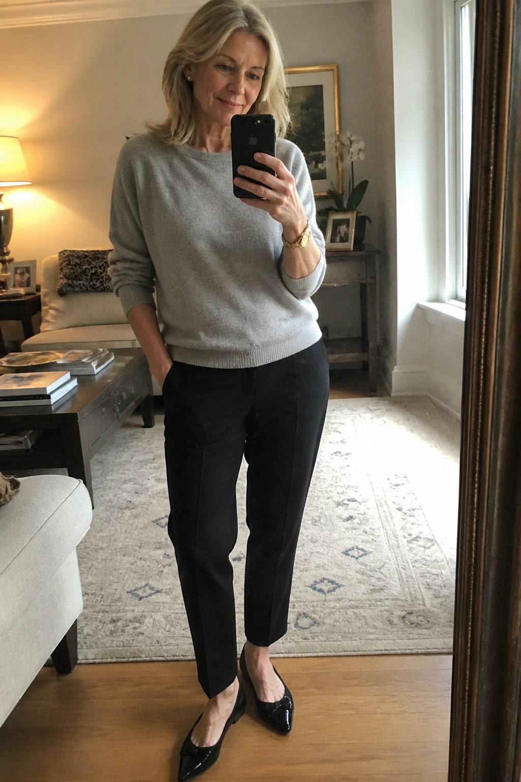 Woman in gray sweater and black pants taking mirror selfie with phone