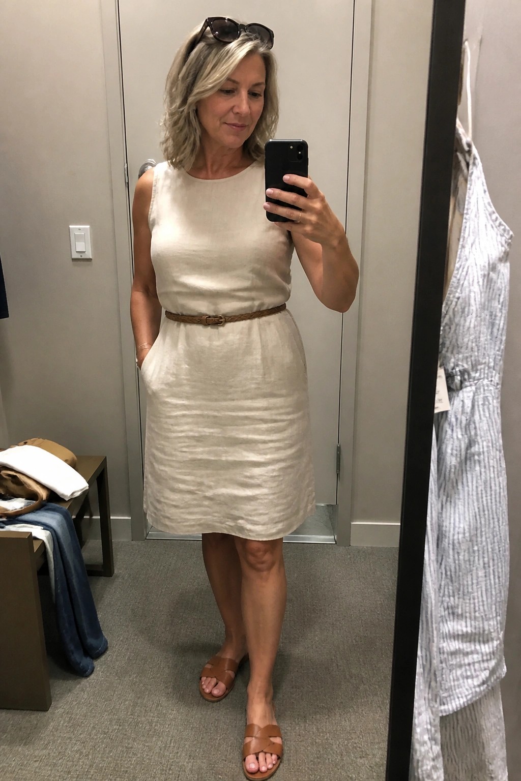 Woman over 50 in cream linen sleeveless knee length dress with brown woven belt and brown slide sandals taking mirror selfie in fitting room