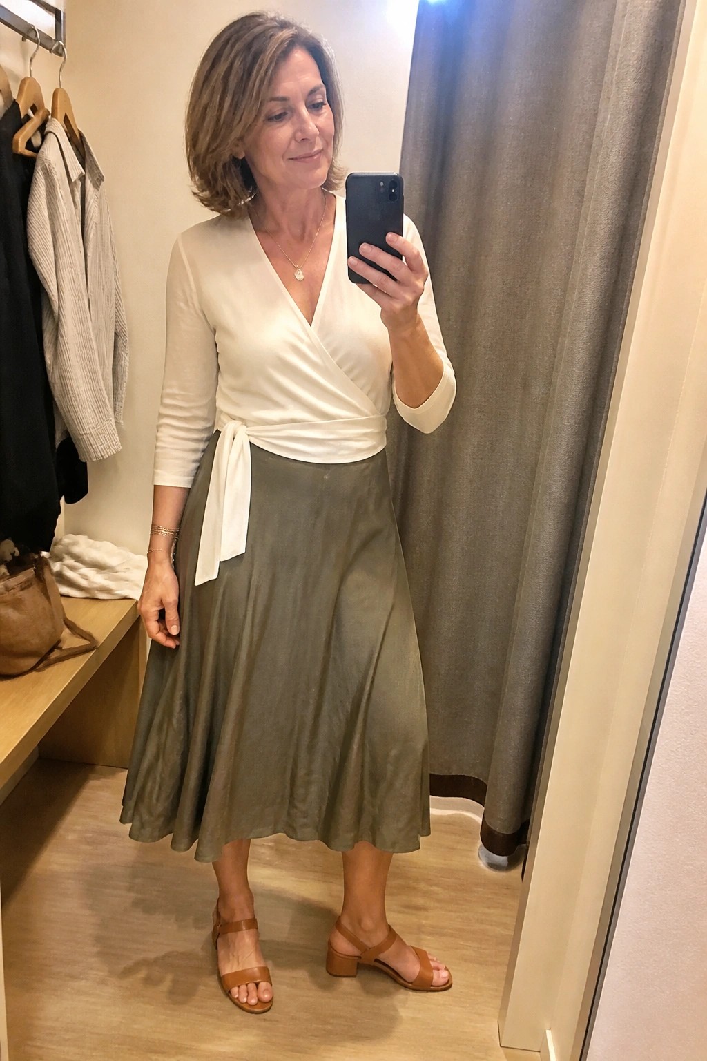 Woman in white wrap top and olive midi skirt taking mirror selfie in dressing room