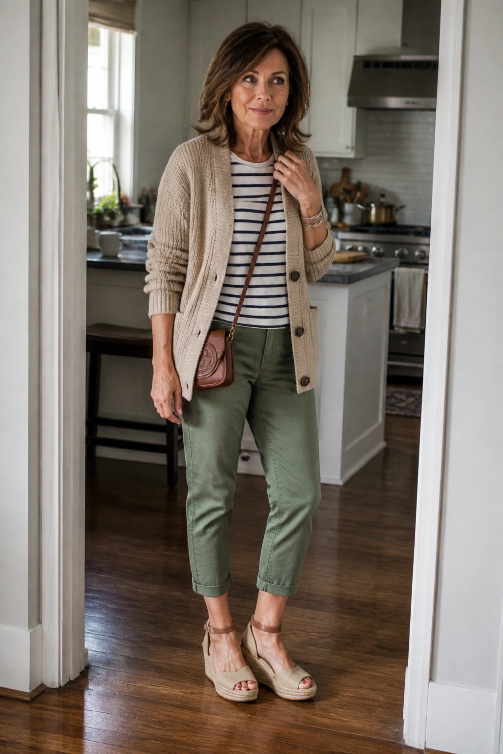 Woman over 50 wearing a beige knit cardigan open over a navy striped tee tucked into olive green cropped pants with a small brown crossbody bag and beige espadrille wedge sandals.