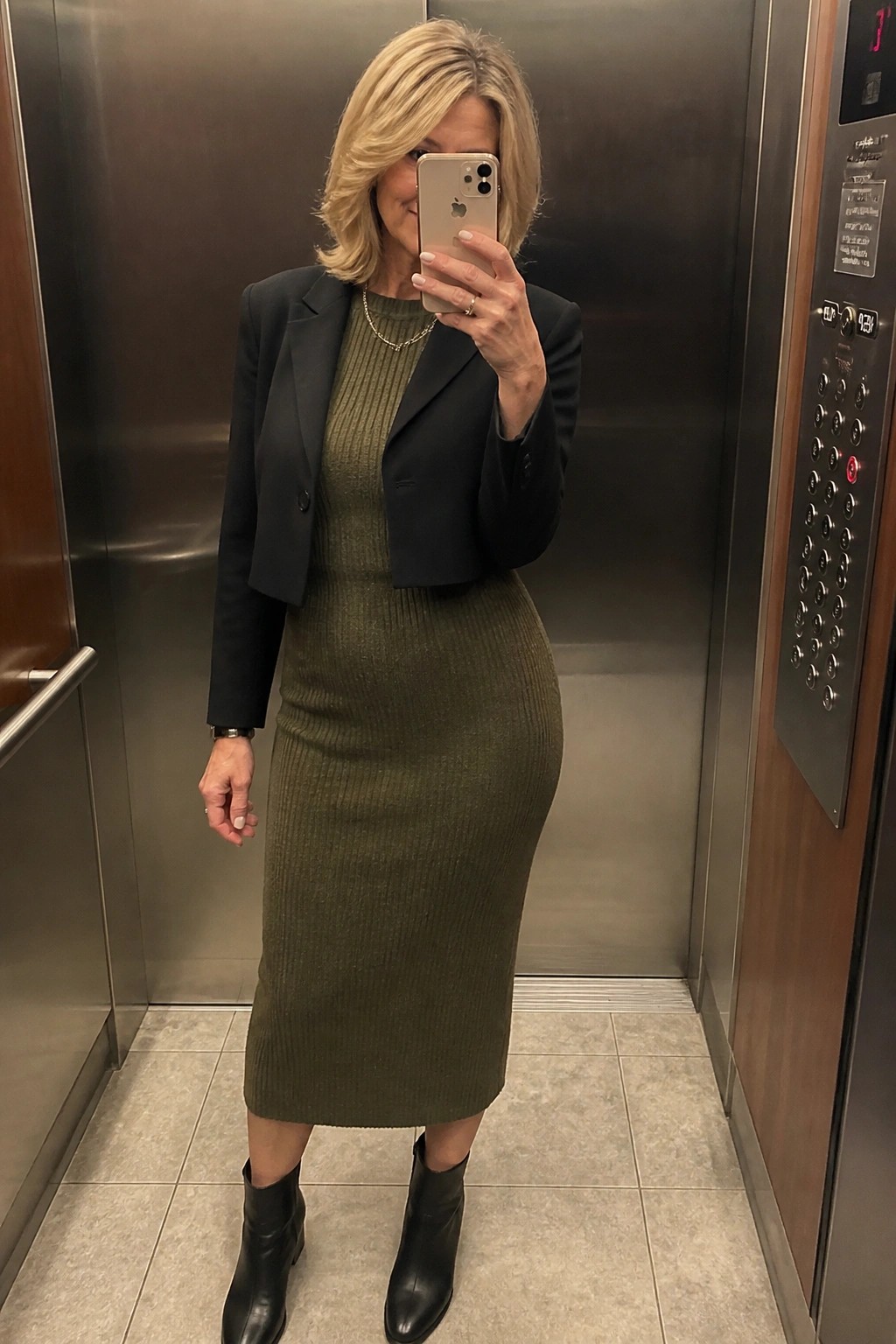 Woman in olive green ribbed dress and black cropped blazer taking selfie in elevator
