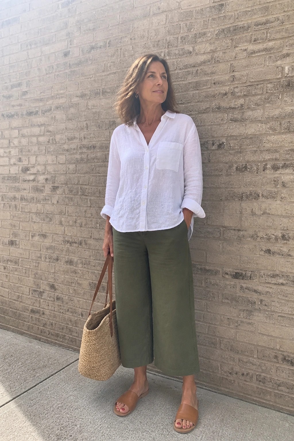 Woman in white linen shirt and green wide-leg pants holding straw bag by brick wall