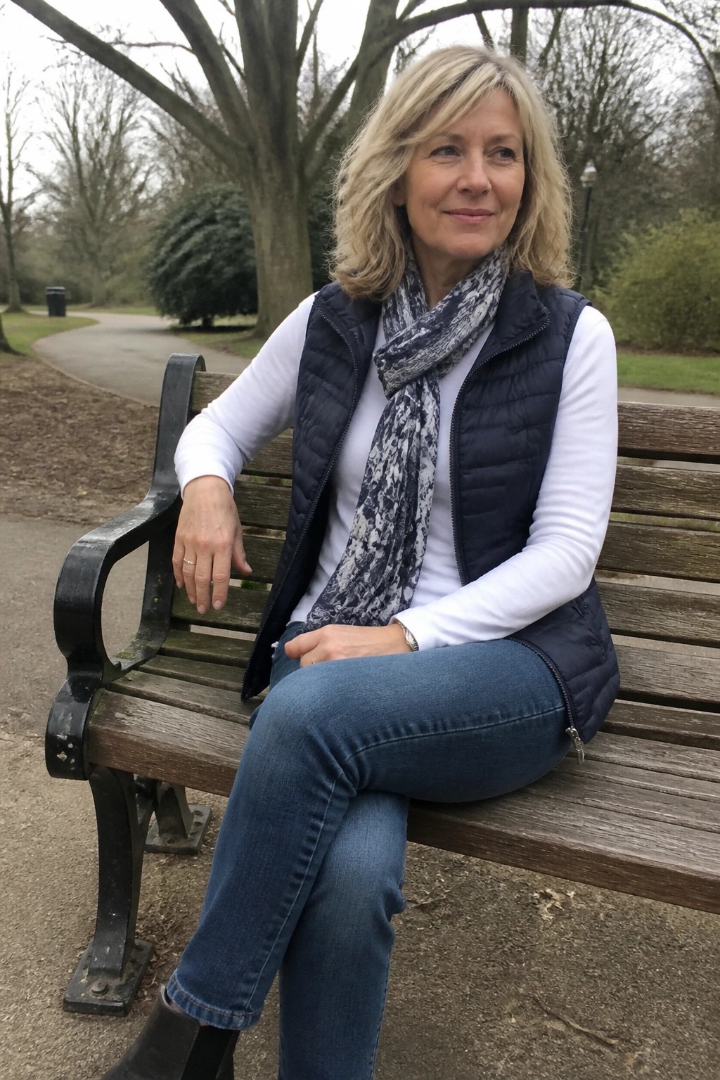 Blonde woman in navy vest, white shirt, and scarf on park bench