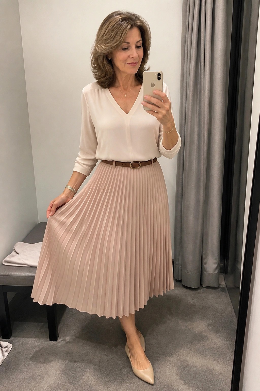 Woman in white blouse and beige pleated skirt taking mirror selfie with phone.