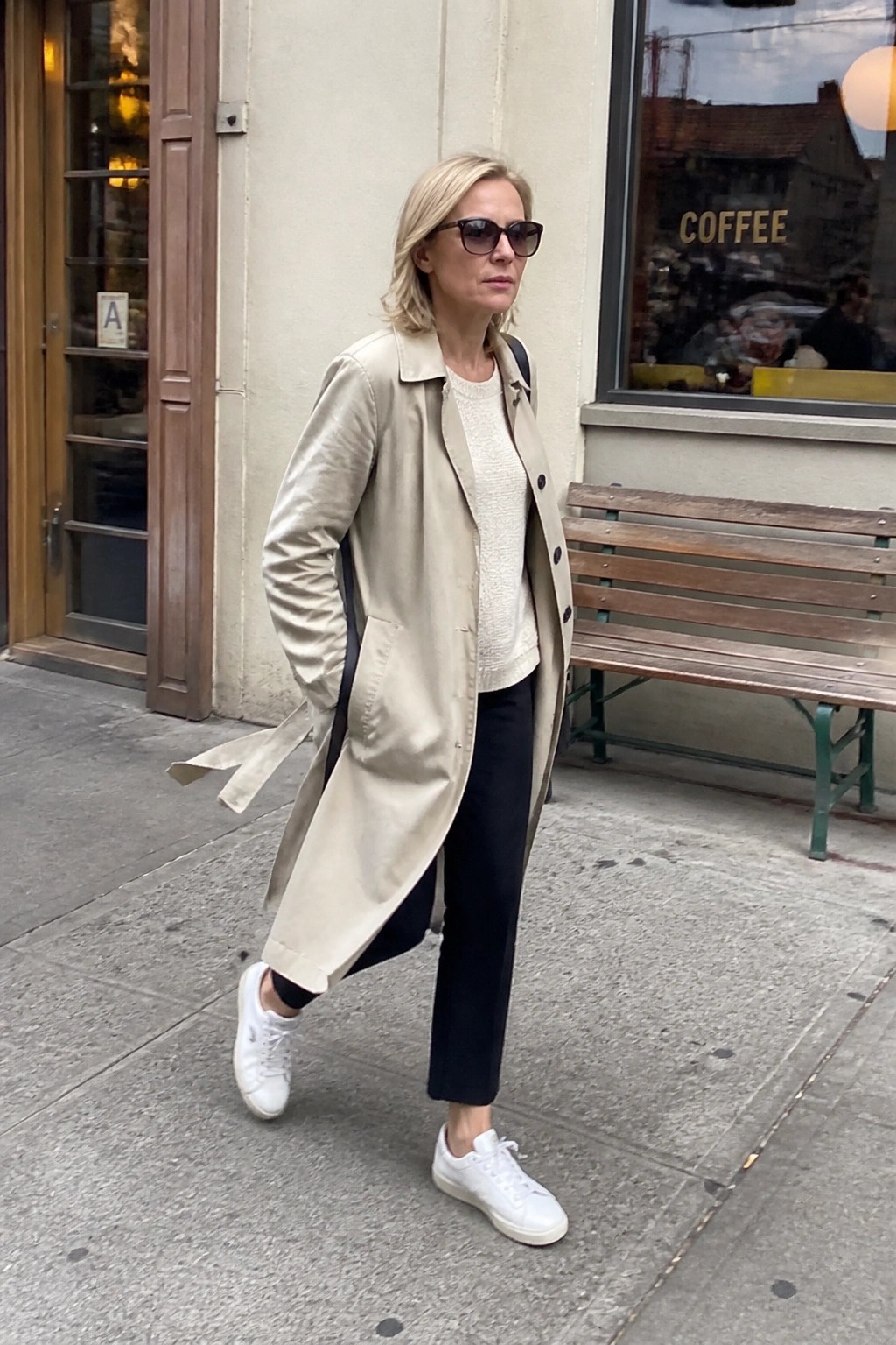 Blonde woman in beige trench coat, white sweater, and black pants on sidewalk
