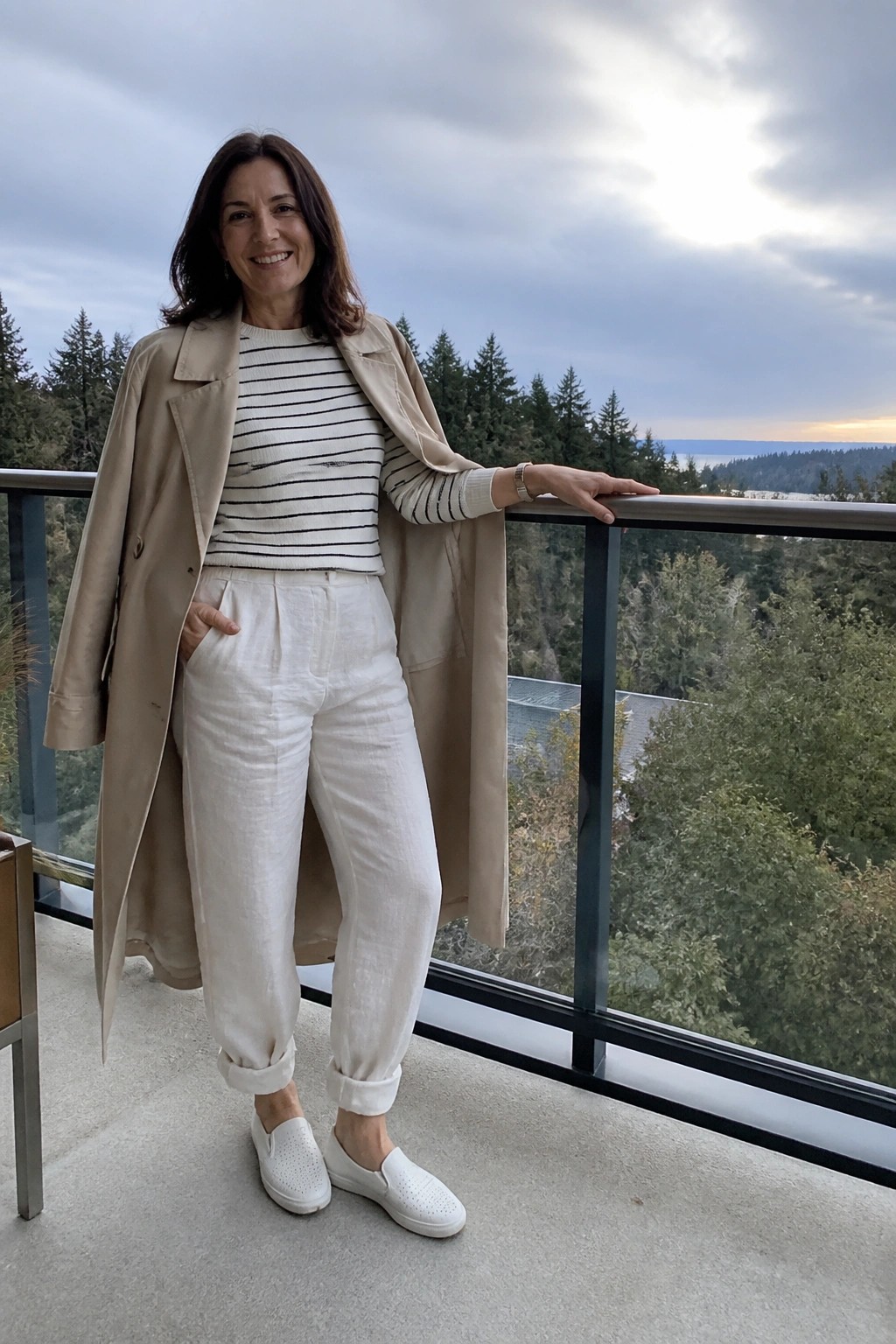 A woman over 50 wearing a beige trench coat over a black and white striped sweater with white cuffed pants and white slip-on sneakers standing outdoors.